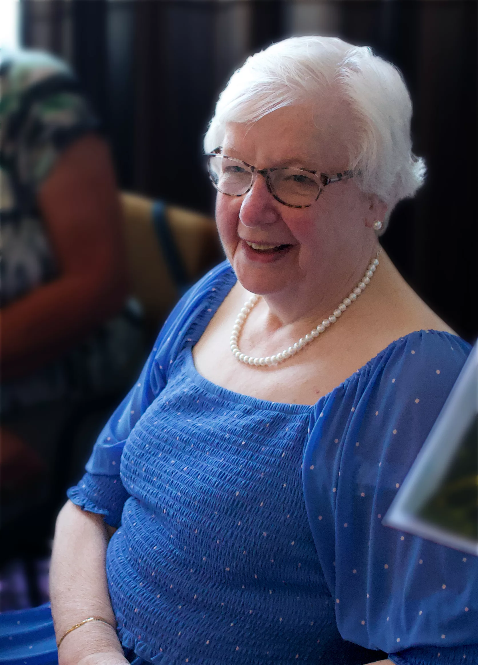 Oudere vrouw met wit haar en een blauwe jurk zit glimlachend te kijken